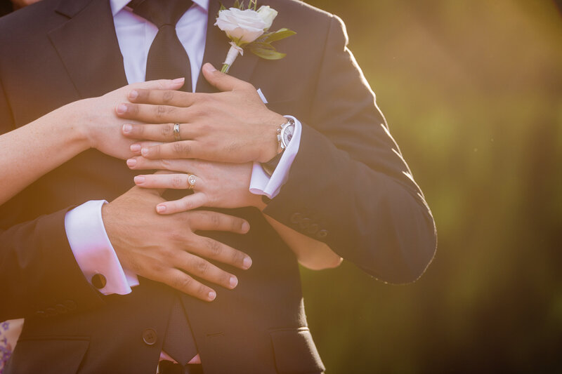 Close-up of the bride’s hands resting on the groom’s chest, showing off their new wedding rings in a heartfelt detail shot.
