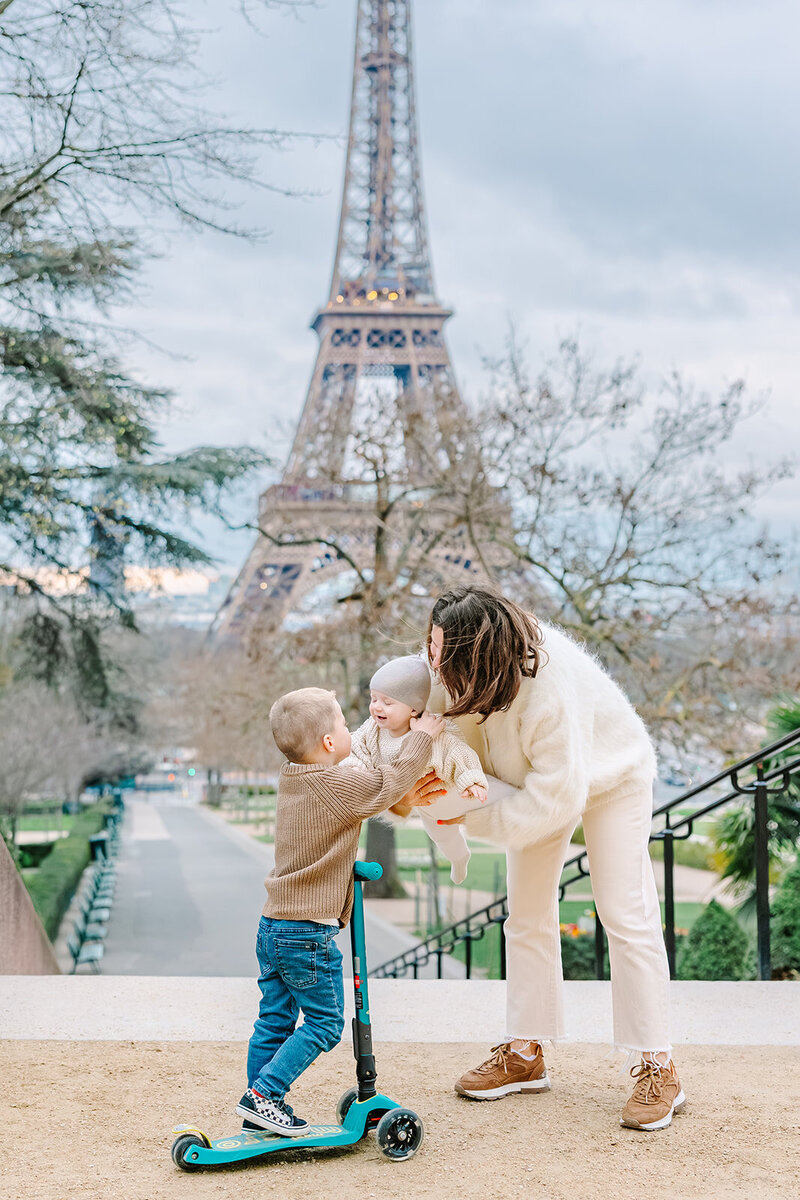 Mother lifting a baby up toward a young boy on a scooter in front of the Eiffel Tower at Trocadéro during a playful Paris family photoshoot.