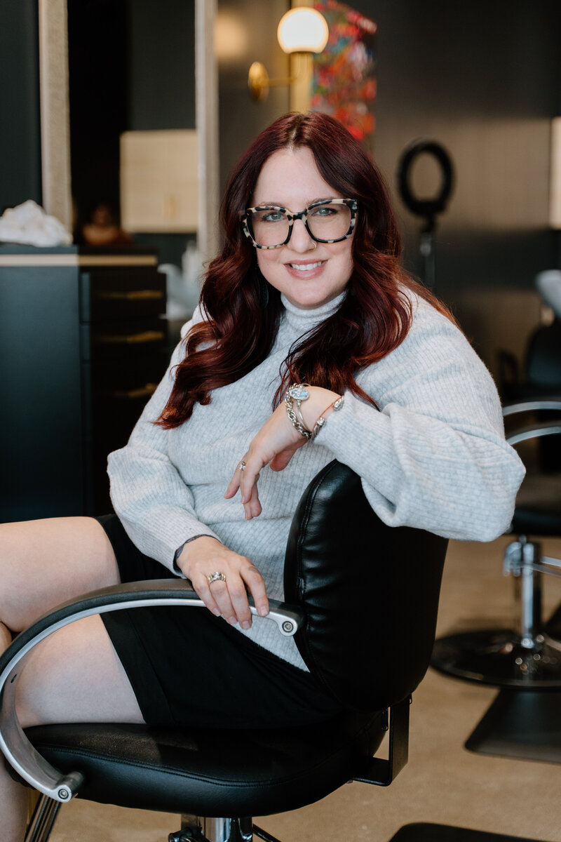 Portrait photography of hair stylist with red hair and tortoise shell glasses sitting in salon chair.