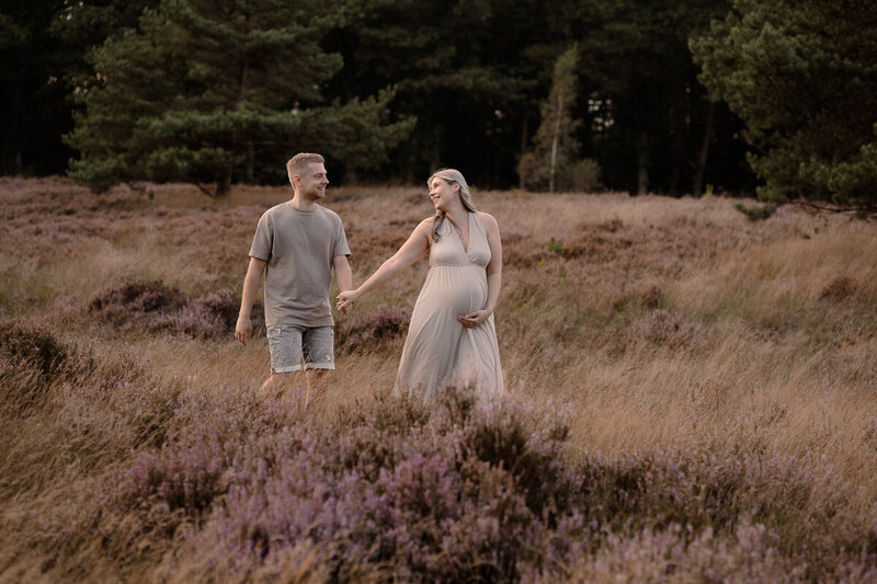 Zwangere dame loopt samen met haar partner over de bloeiende heide tijdens een zwangerschapsshoot