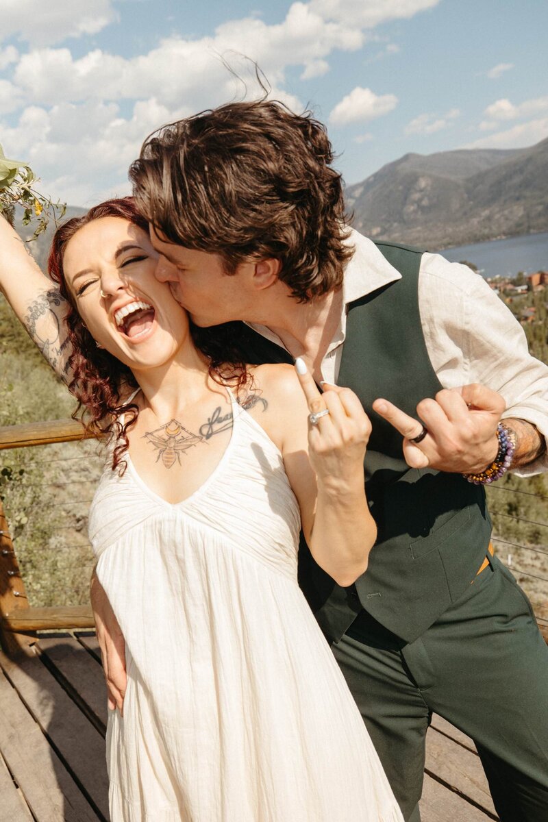 A bride and groom stand in front of a mountain lake, both holding up their ring fingers to the camera. The bride raises her bouquet in the other hand, and the groom kisses her on the cheek.