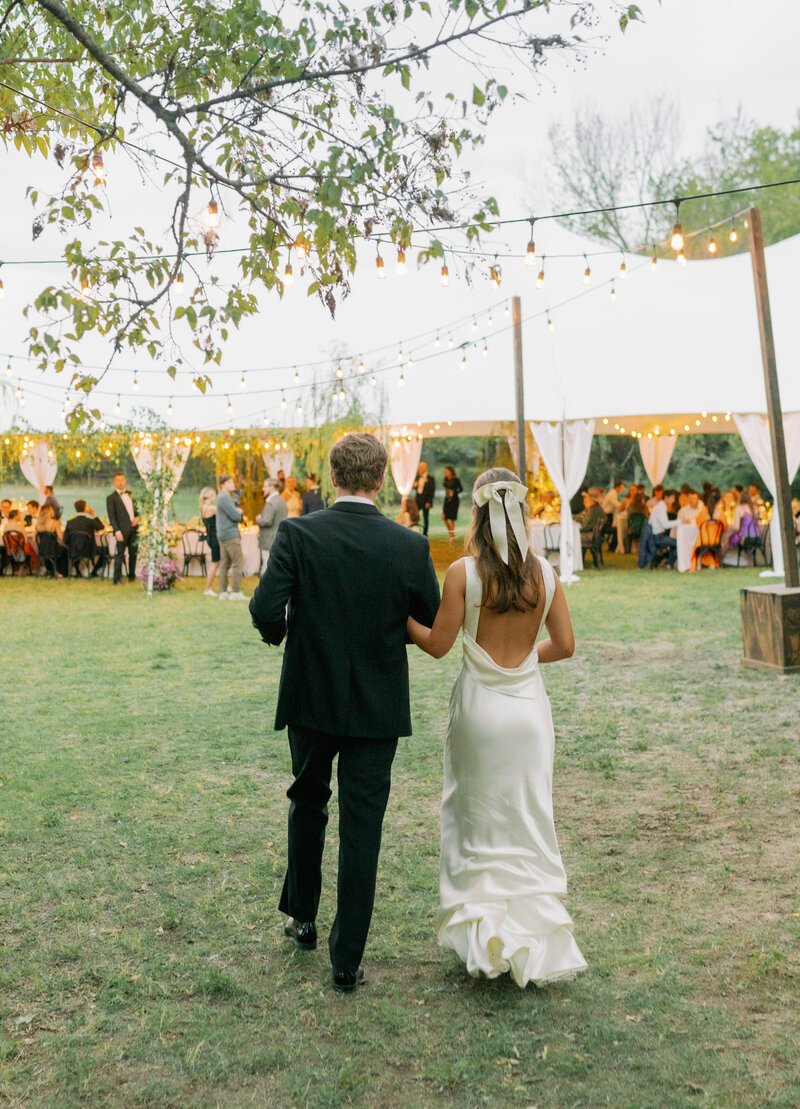 Bride holding groom's arm while they walk towards their outdoor reception tent under string lights