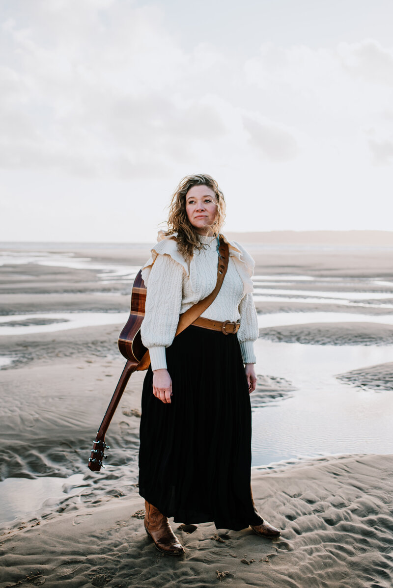 woman with a guitar on her shoulder is stood confidently on a low tide beach as the wind blows her hair