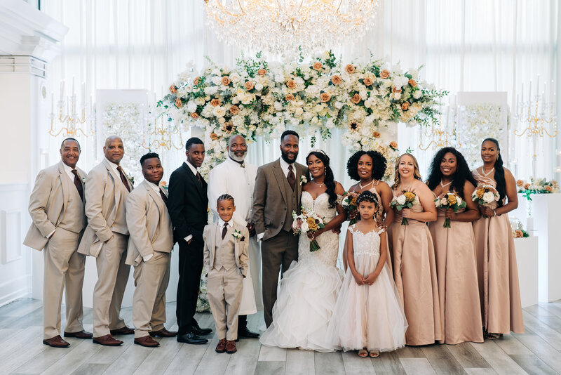 a wedding party poses in front of the ceremony space with chandeliers and florals behind them