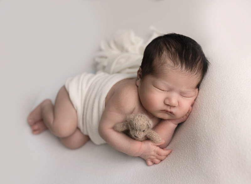 baby boy laying on his side, on a beige backdrop and holding a teddy bear