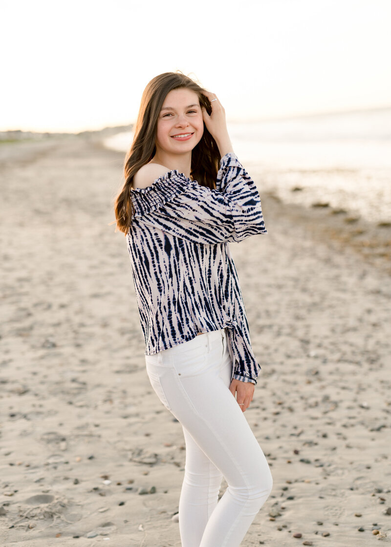girl standing on the beach touching her hair wearing a blue and white shirt and white jeans