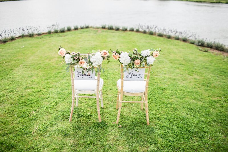 Two light wooden chairs decorated with white and peach flowers and greenery, placed on a grassy lawn facing a body of water. The chairs have signs that read 'Forever' and 'Always' on the backs