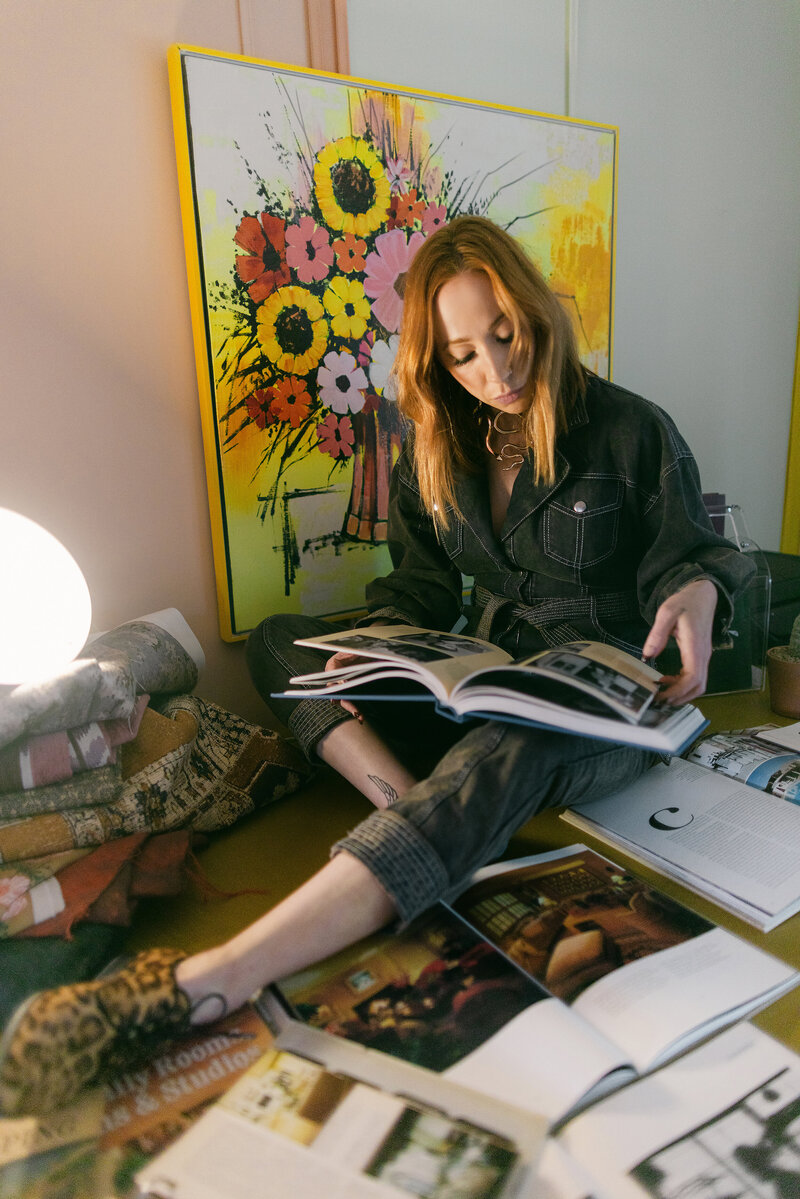 Artistic branding photo of a woman reading surrounded by design books and vibrant floral artwork, captured in a retro-modern studio by Dani Lacey Photographs in Greenville SC.