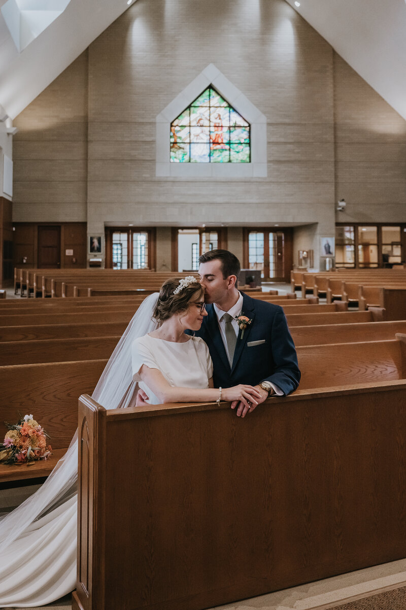 groom kissing bride on the head while they sit on a church pew