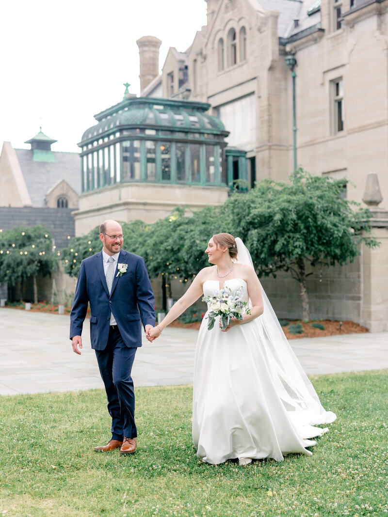 Bride and groom holding hands and walking outside of a large stone wedding venue