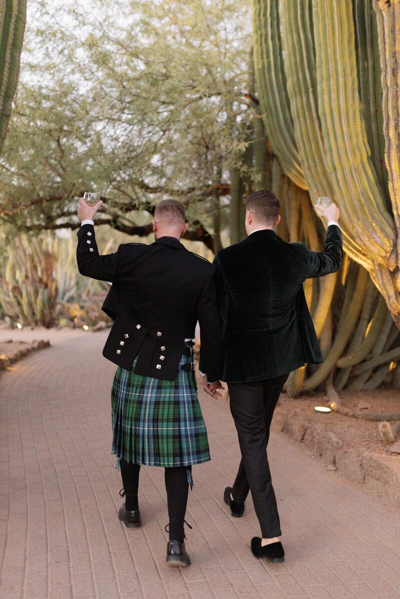 Two grooms walk away holding hands in a cactus garden and cheers their cocktails together.