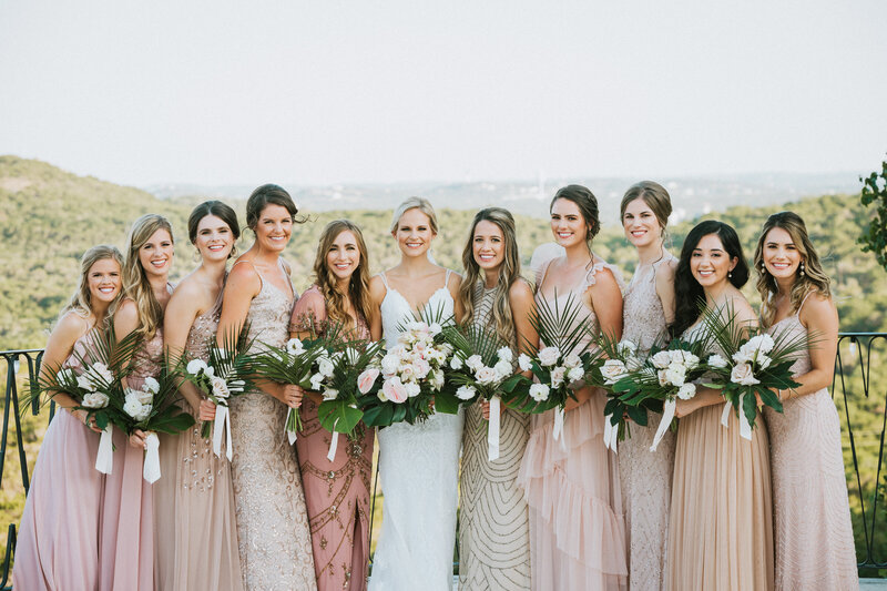 Bride and bridesmaids wearing different dresses in complementary tones, posing together with a stunning view of the Texas Hill Country at Villa Antonia.