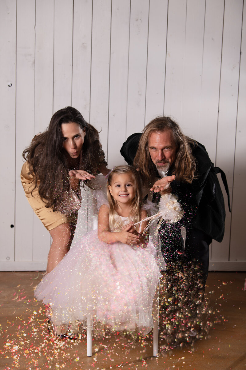 Glittery Family Portrait for New Year's. Mom and Dad pretending to blow glitter in front of their daughter. Photo taken by Lydia Teague Photography.