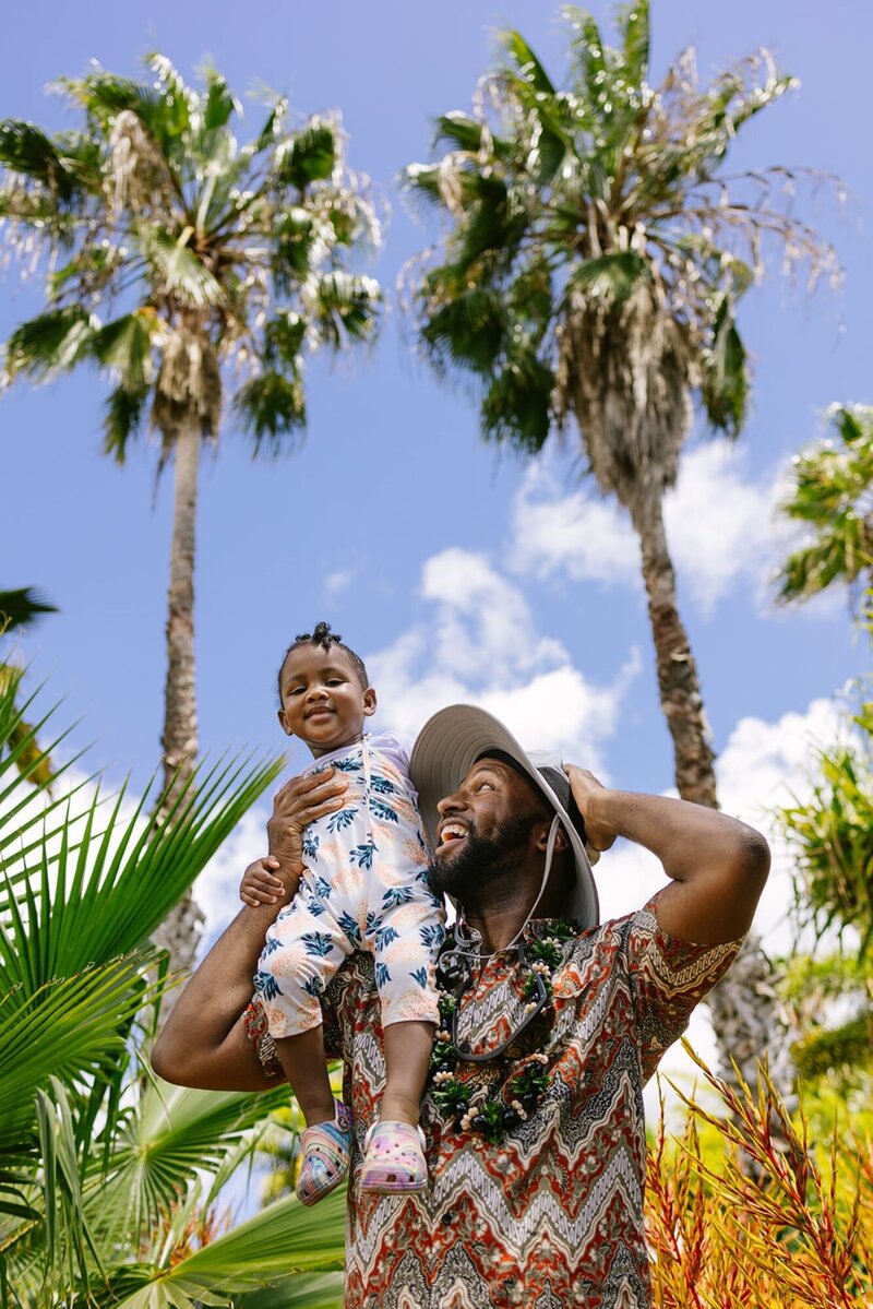 A man hold his toddler on his shoulder with palm trees shown up in the sky.