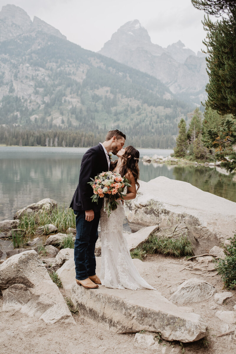 outdoor fall wedding with bride adn groom kissing as they stand on a large boulder on a lake in the mountains with  the grand tetons in the distnace captured by jackson hole photographers for a wyoming elopement