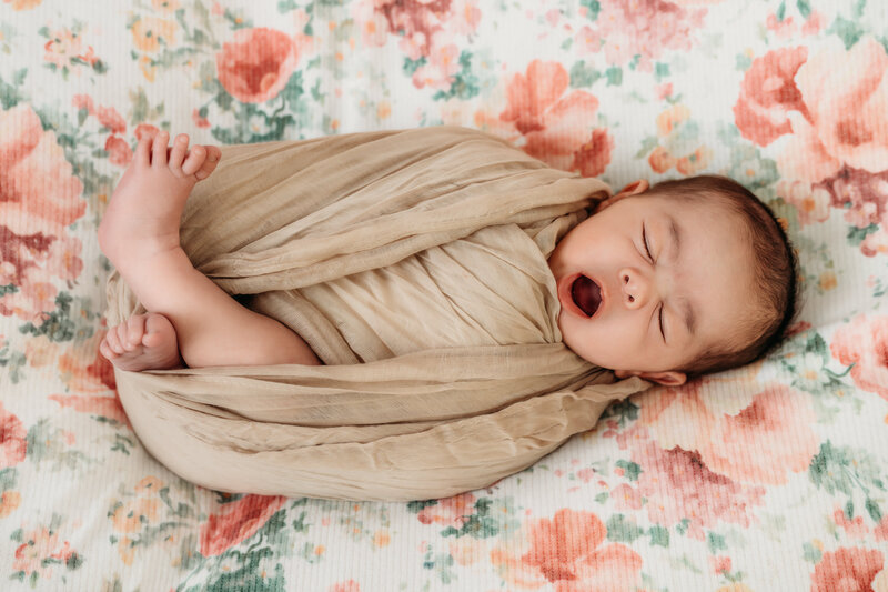 newborn wrapped in tan fabric during her studio session. she is yawning and you can see her toes sticking out of the wrap.