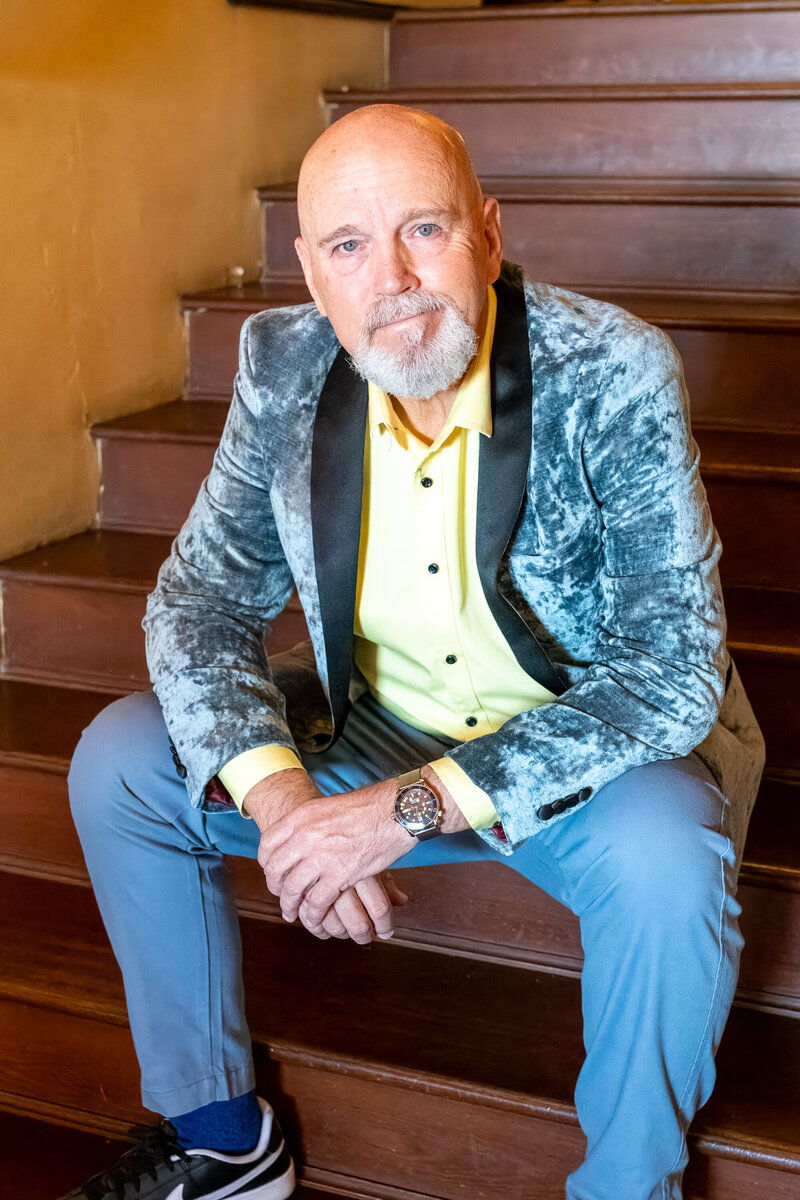 A man seated on a wooden staircase wearing a velvet blazer and yellow shirt, photographed by Vyrl Photo as part of Tucson brand photography.