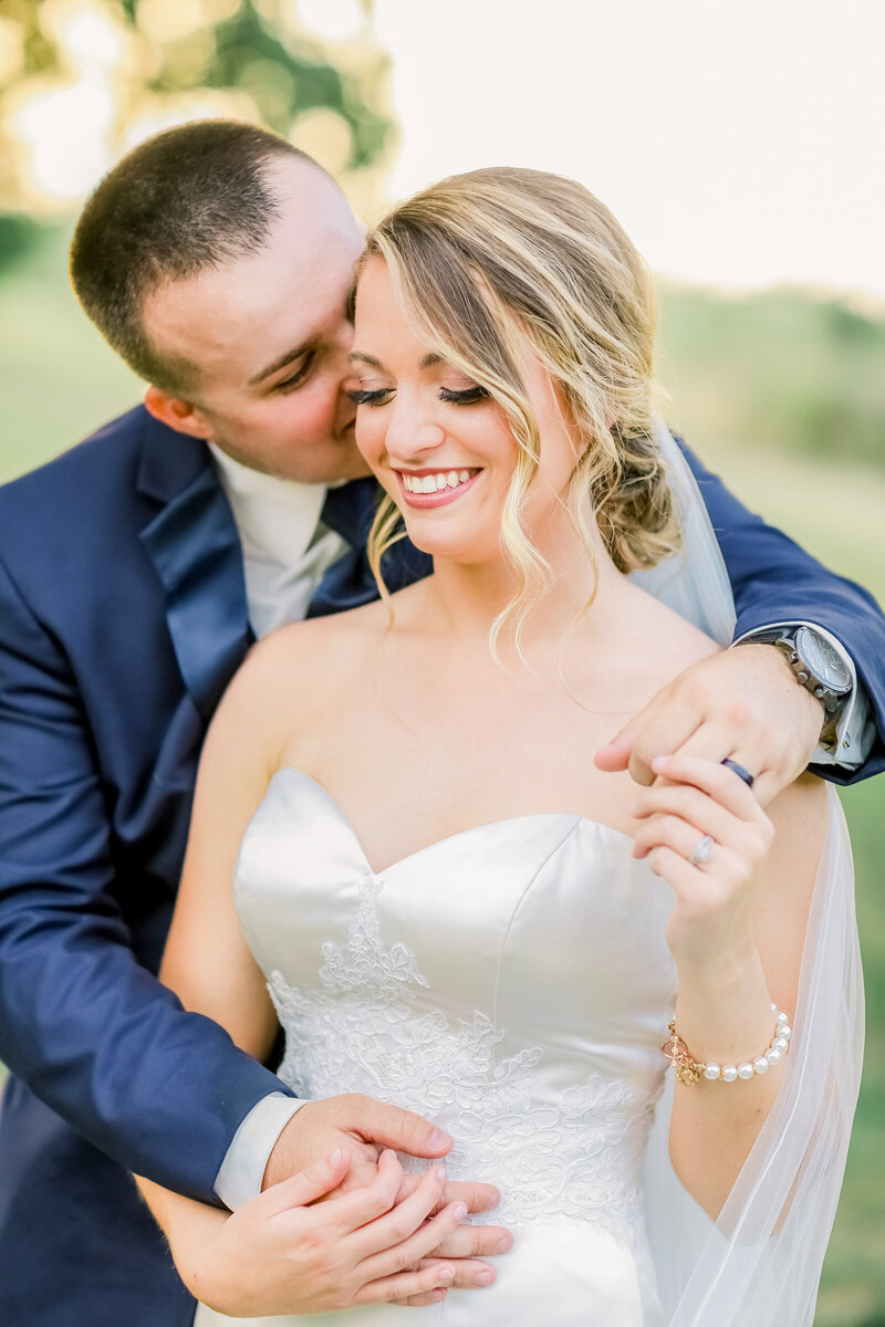bride-and-groom-smiling-holding-hands-groom-kissing-brides-cheek-bride-with-pearl-bracelet-bride-wearing-satin-and-lace-wedding-dress-groom-wearing-navy-blue-suit-wedding-at-mighty-oak-lodge-in-lebanon-mo-by-branson-wedding-photographer-kathryn-faye-photography