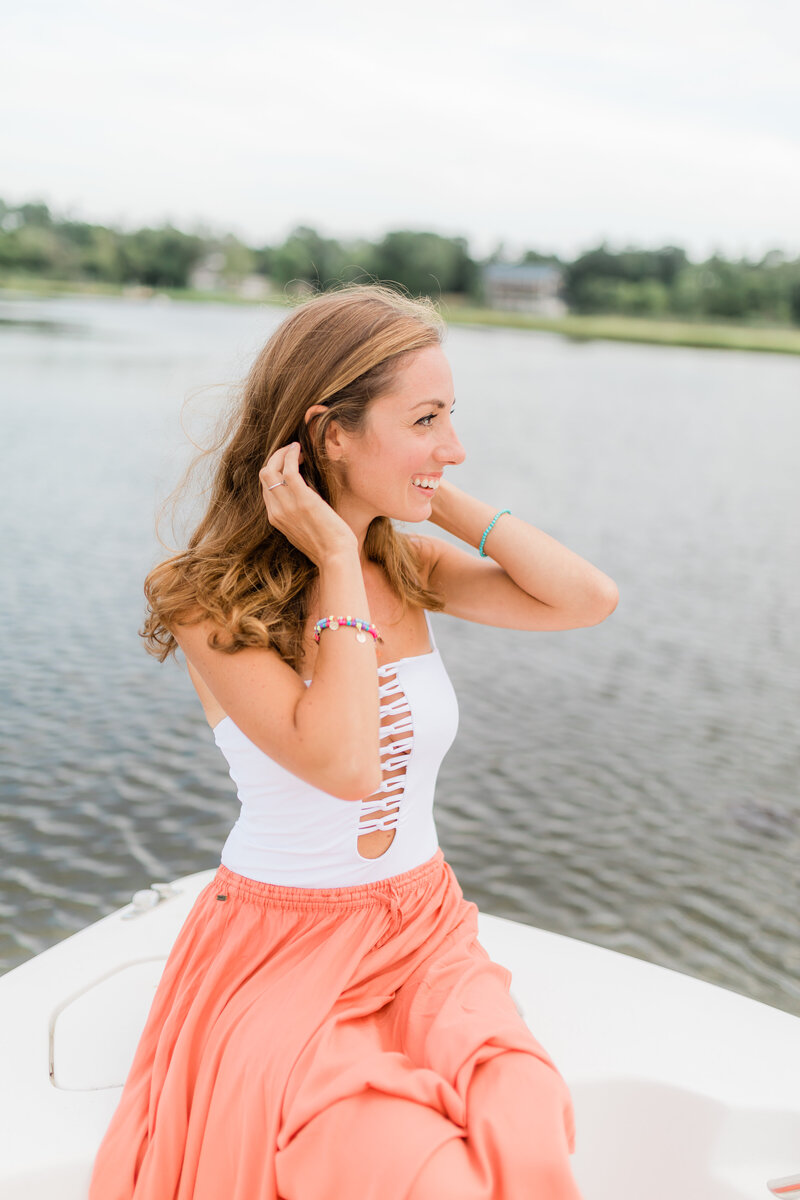 Best senior photographer in New Jersey. Portrait of a photographer, sitting on a boat. She is smiling and looking to the right while holding her hair back from the wind. She is wearing an orange maxi skirt and white bathing suit with beaded jewelry.
