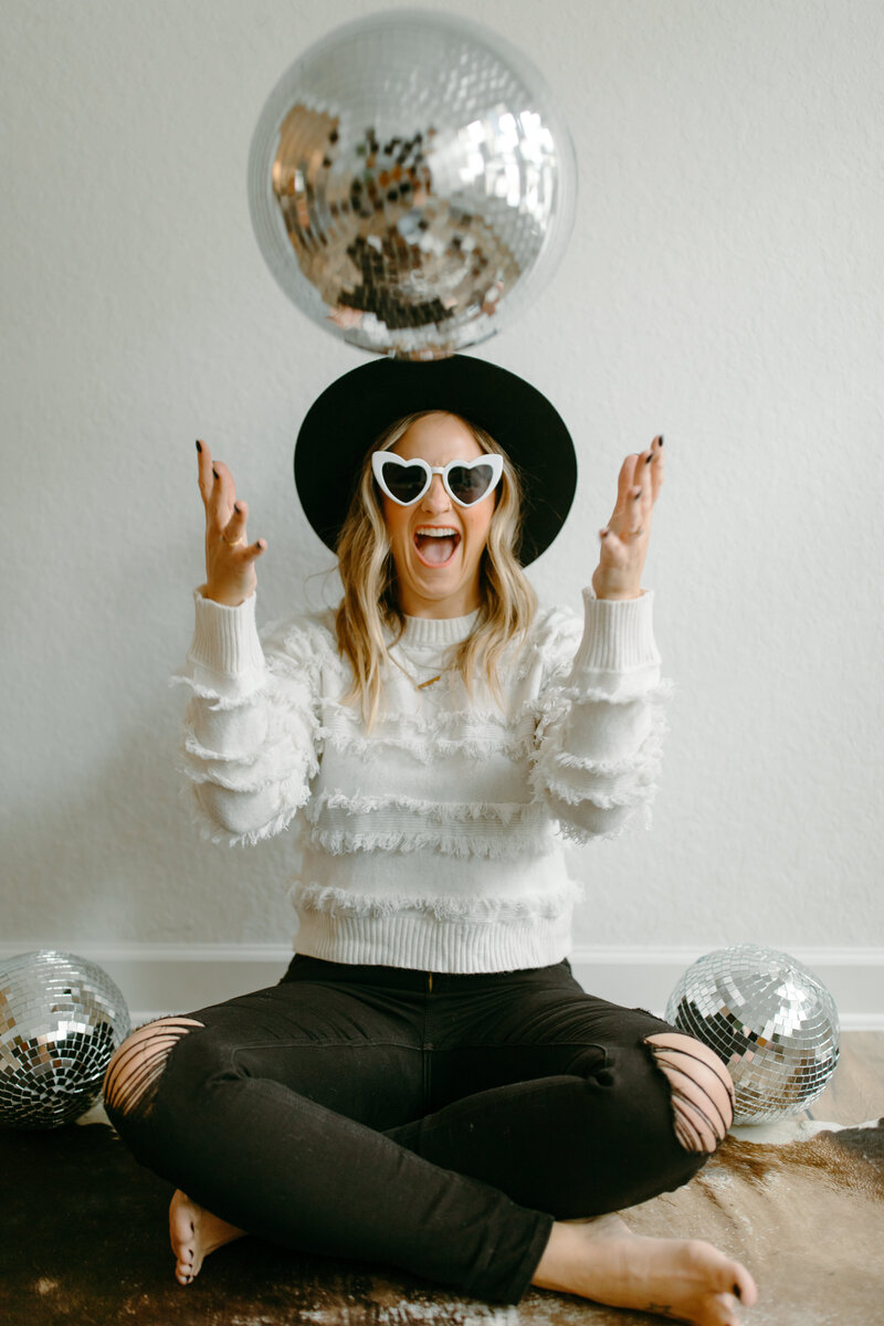 Jessie Schultz throwing up a disco ball with heart sunglasses on  and a huge happy smile on her face. Disco balls around her sitting on the floor.