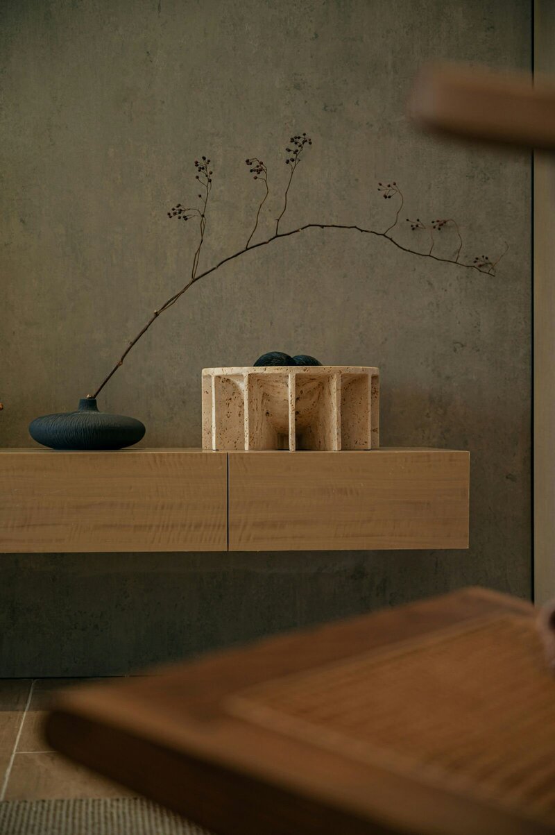 A serene and sophisticated unhurried interior vignette, showcasing a floating oak cabinet set against a textured plaster wall. The arrangement features a dark, organic vase with a delicate branch and a sculptural stone object, highlighting natural materials, subtle textures, and the enduring beauty of deliberate design.