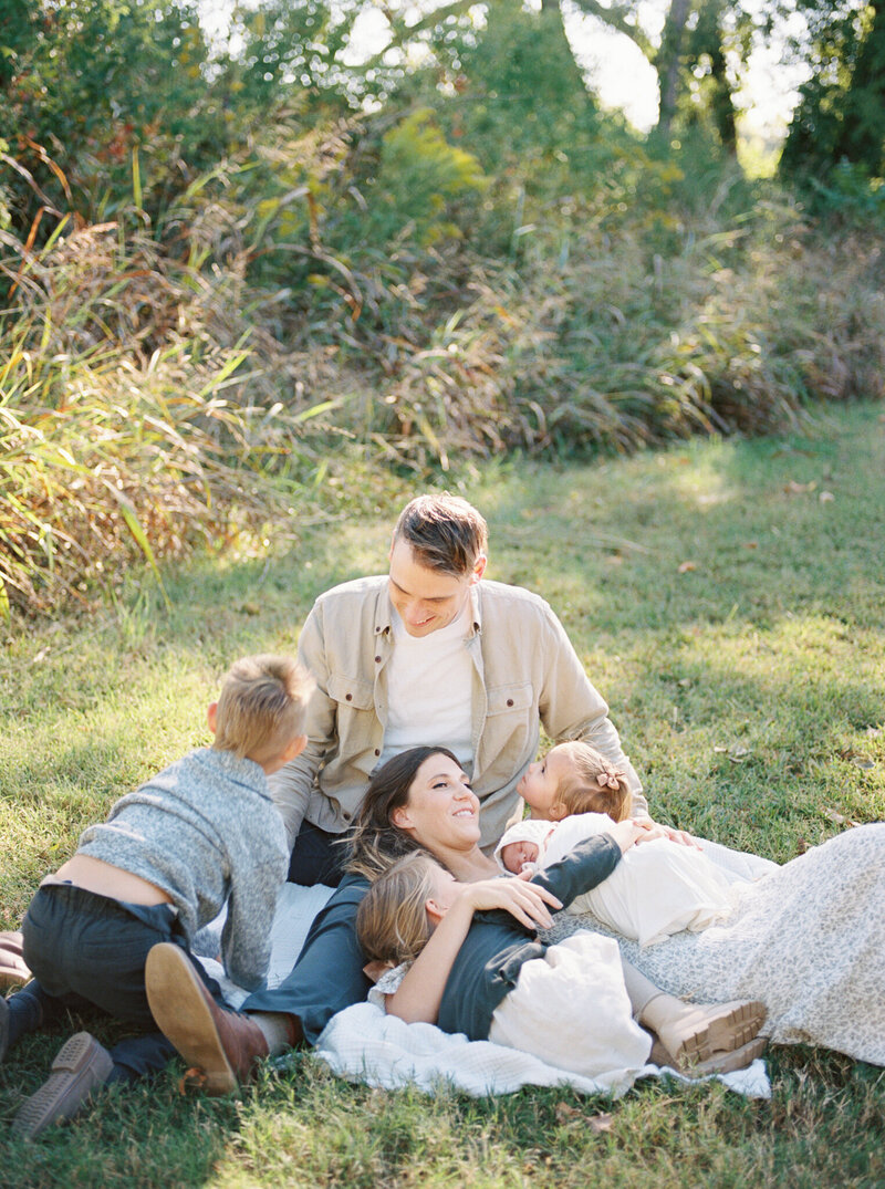 Mother lays down on her husband's lap amidst grass while her 4 children snuggle each other