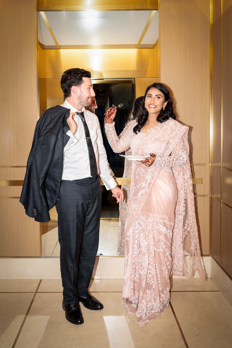 A man in a suit holds his jacket over his shoulder, smiling at a woman in a pink lace saree. She is smiling back, holding a slice of cake. Theyre standing in an elegant room with wooden paneling.