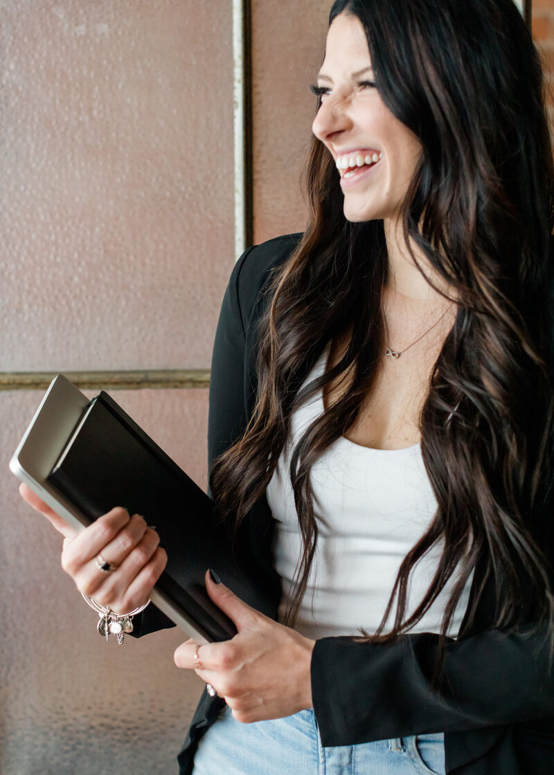Allie Clark smiling while holding a notebook, dressed in black blazer and white top, standing against a window.