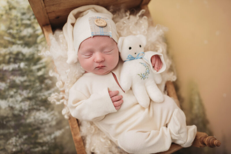 newborn baby boy in white outfit and white sleepy cap laying with teddy bear in tiny wooden bed prop with winter trees theme backdrop