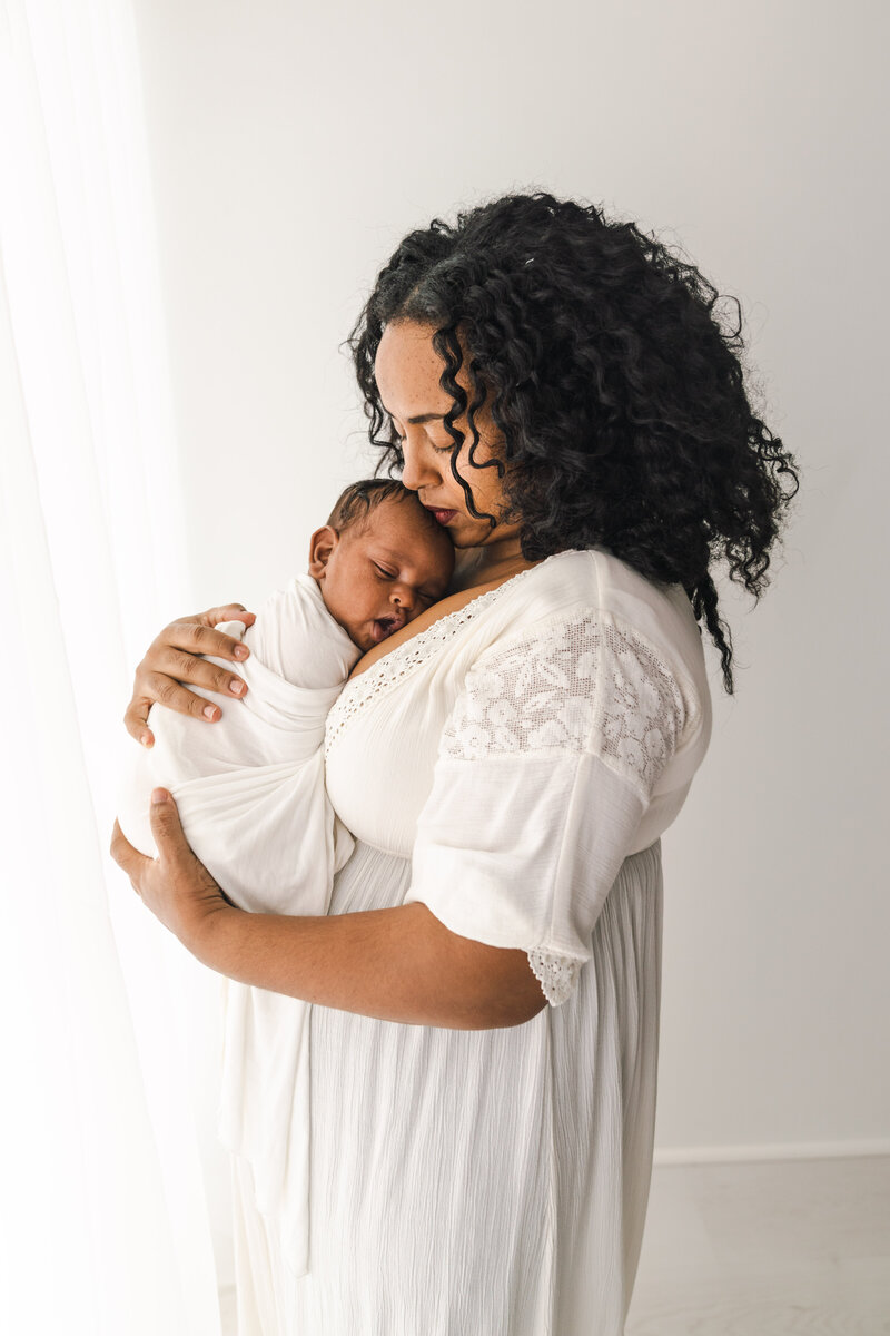 A Minneapolis newborn photographer captures a woman gently cradling a sleeping baby in a serene, white-lit room.
