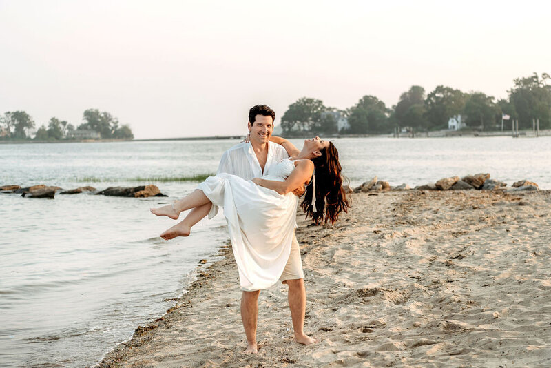 Partner carries expecting mother in flowing white dress along Connecticut beach during romantic waterfront maternity session with Connecticut maternity photographer. Couple poses barefoot at water's edge during golden hour