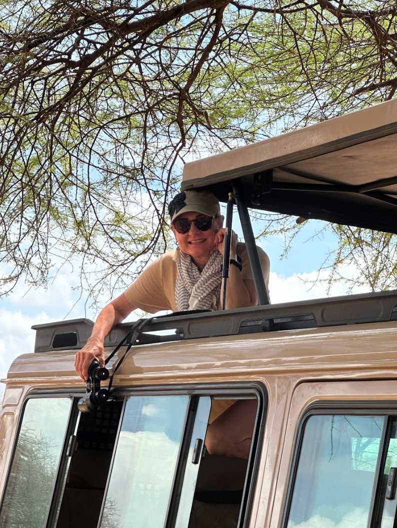 A woman wearing sunglasses, a cap, and a beige shirt, leans out of a safari vehicle with a camera, smiling, ready to capture wildlife. The vehicle’s roof is popped open for better visibility, with the African wilderness in the background.