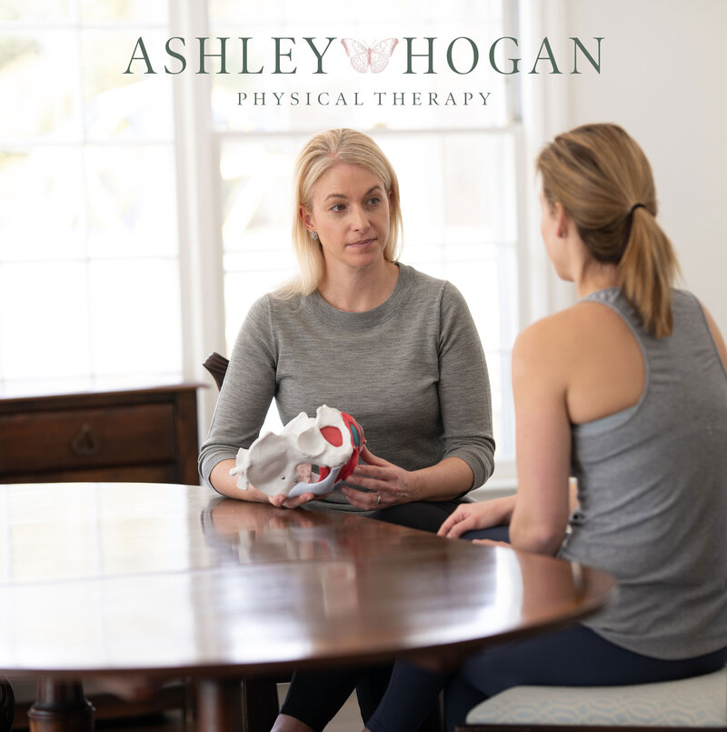 Two women discuss treatment while sitting at wooden table with Ashley Hogan Physical Therapy logo centered at top