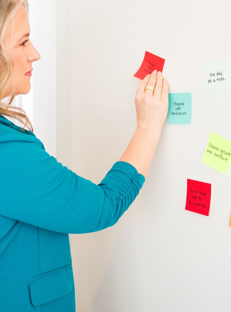 Woman in a teal blazer putting sticky notes with inspirational messages on them on the wall.
