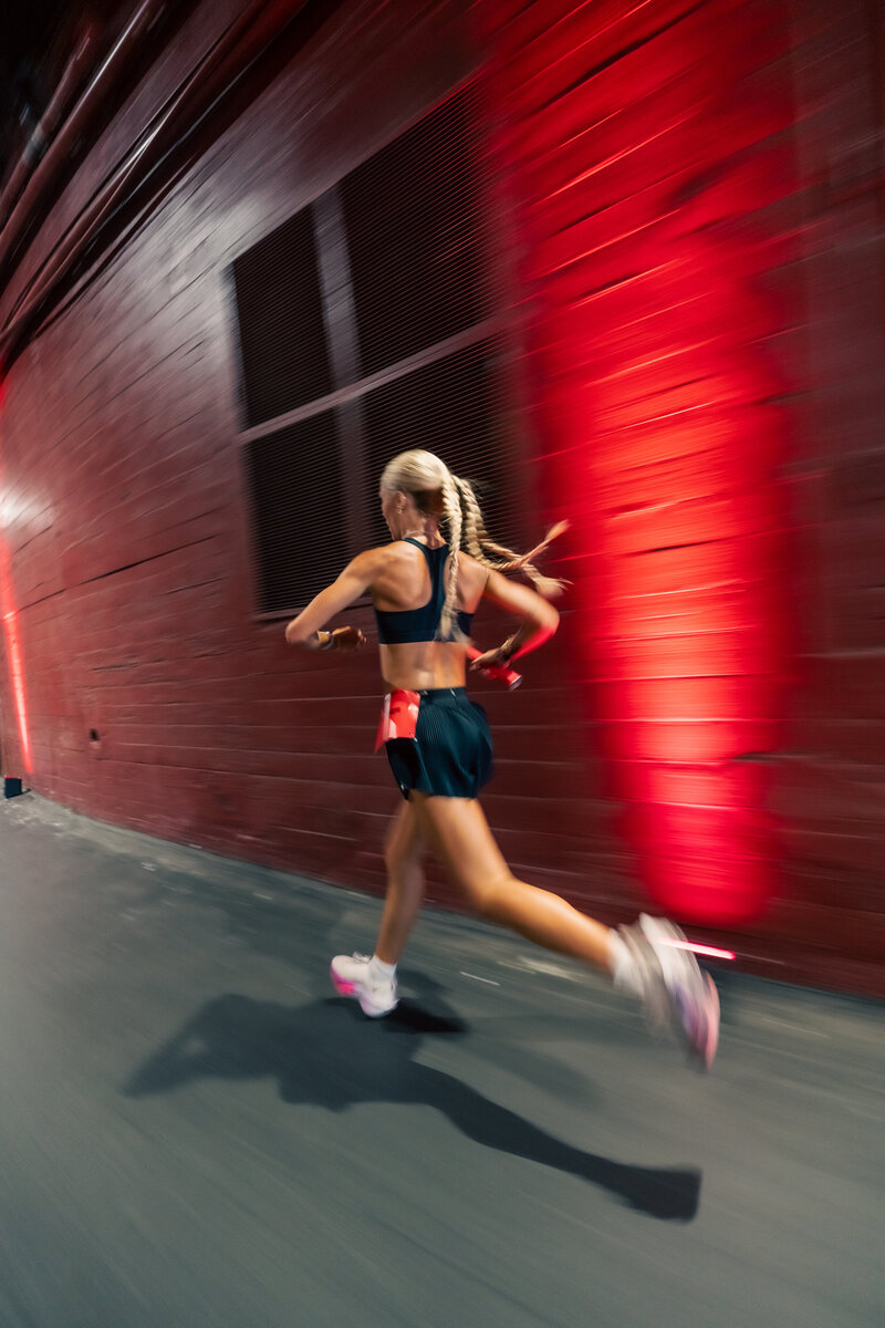 A HER Sports athlete sprinting past a red wall during training, holding a relay baton while running at a fast pace.