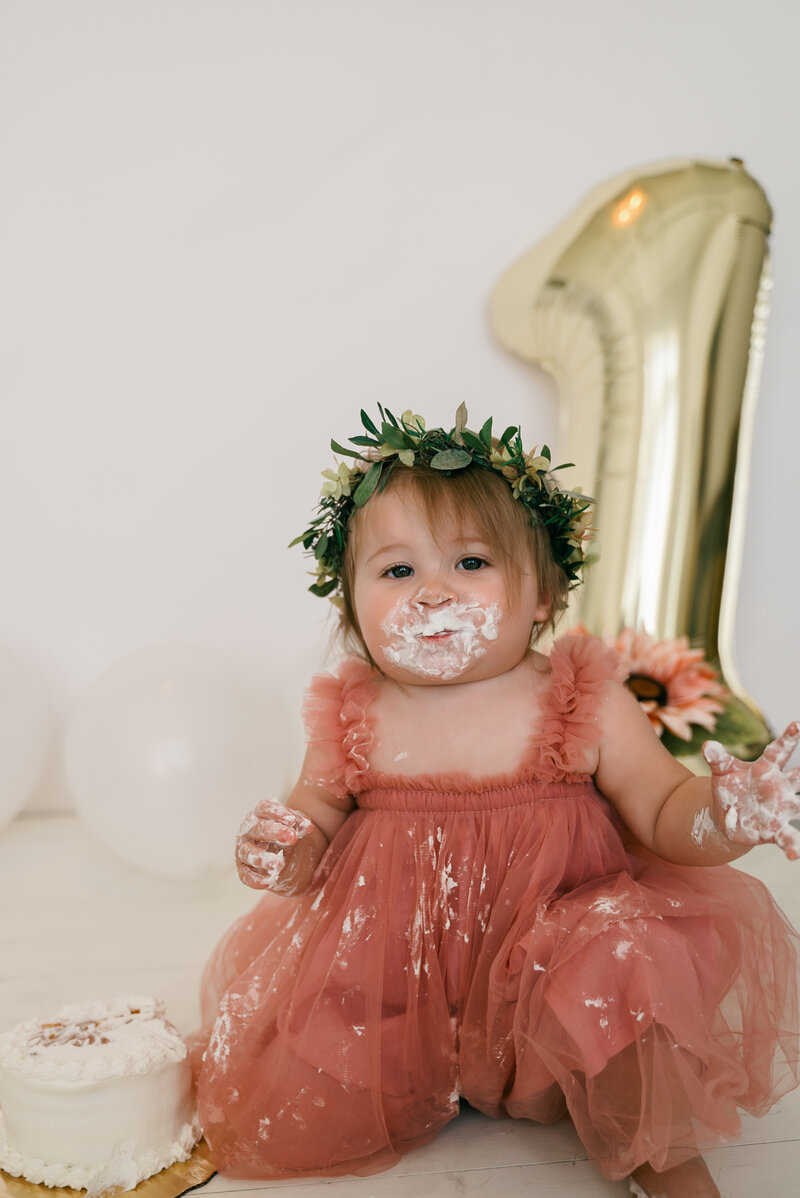 Baby girl in pink dress and leaf headband covered in icing from birthday cake.