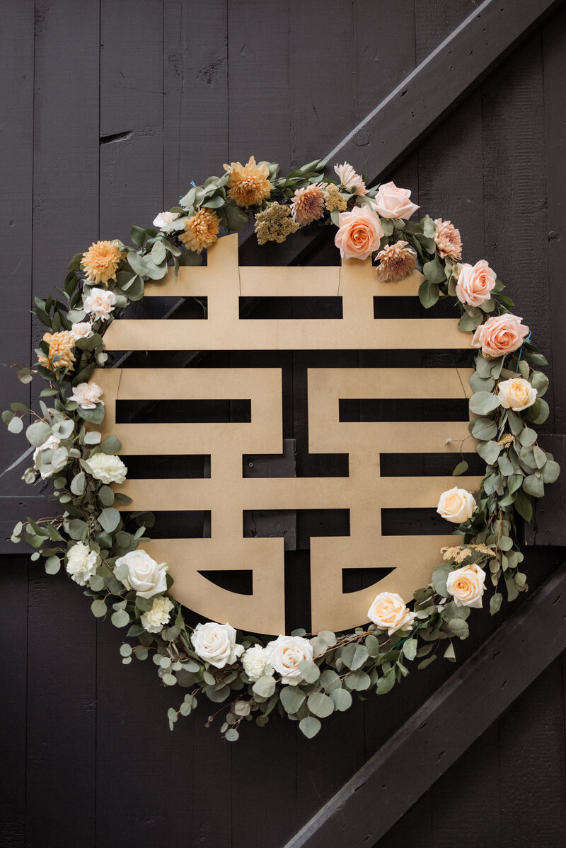 A pastel floral wreath hangs on a black barn door. Inside the wreath there is a gold Chinese character that reads "double happiness"