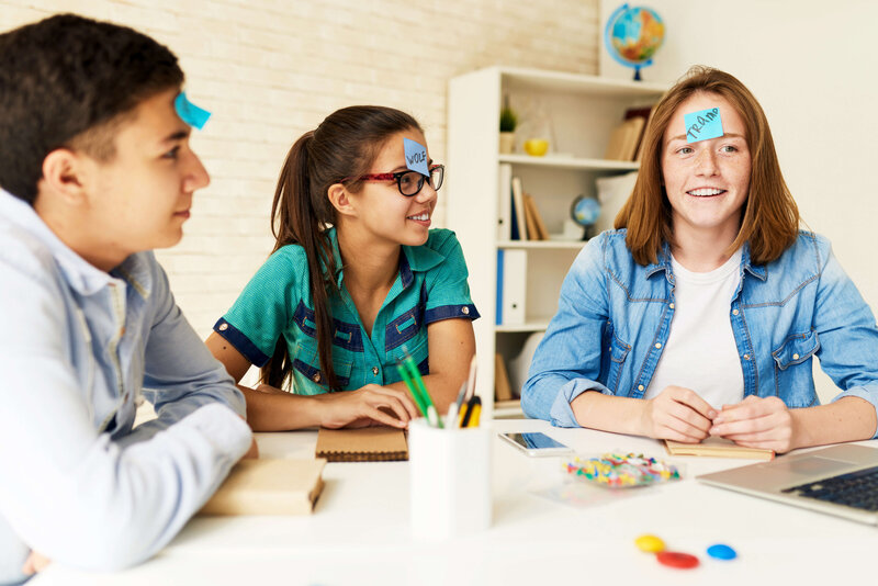 three kids at a desk with blue sticky notes on their foreheads smiling