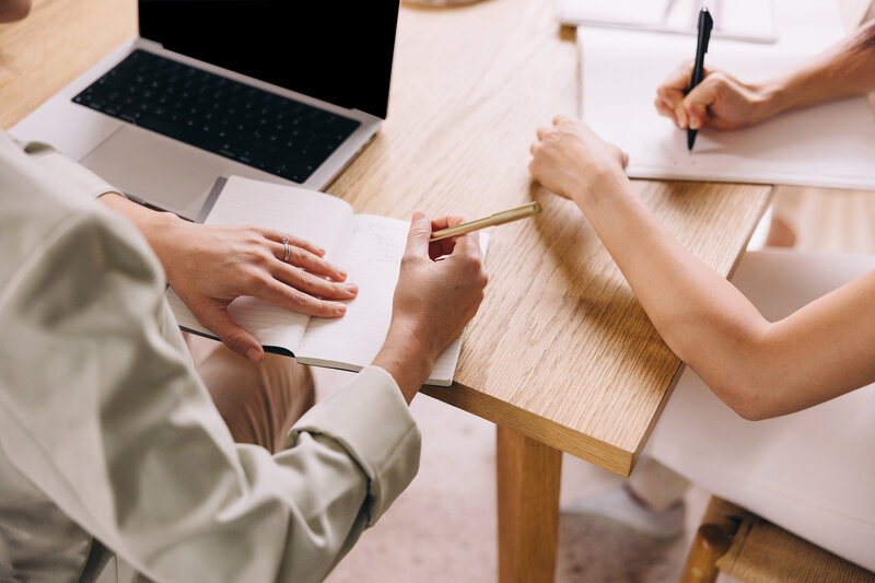 Two people collaborating at a light wood desk, taking notes in open notebooks during a meeting, with a laptop nearby. Captures the clarity and momentum gained from a 60-minute follow-up call to review a custom business roadmap and next steps.