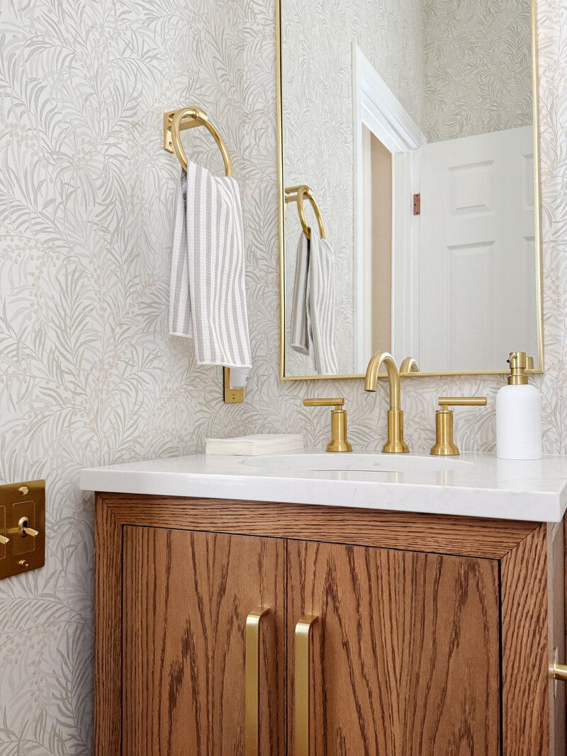 Custom vanity with brass hardware, neutral patterned wallpaper, and a tan and white striped towel