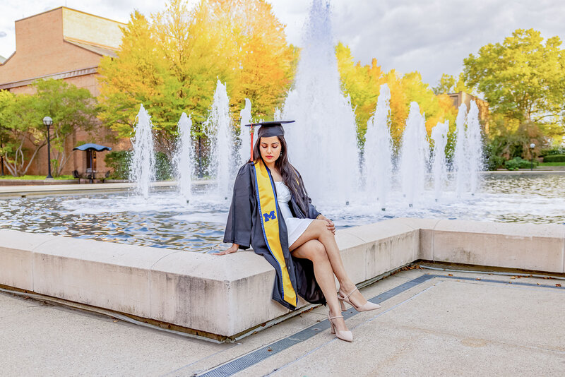 A female University of Michigan graduate is sitting in her cap & gown next to the Lurie fountain. The woman is looking down at her hand