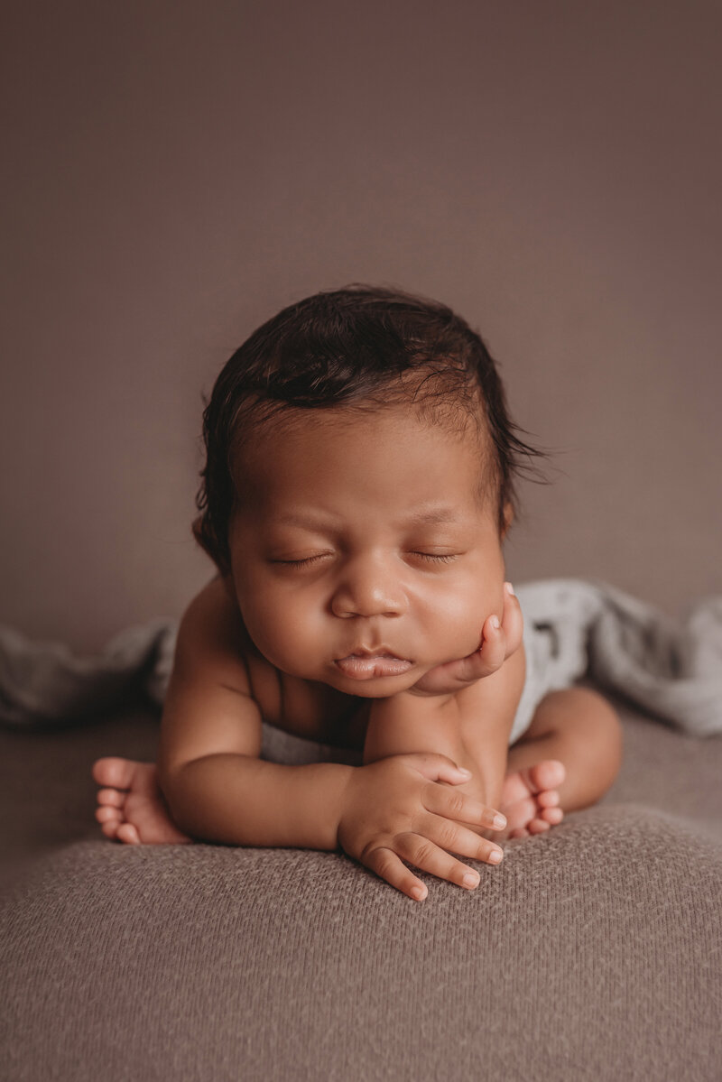 Newborn baby boy asleep posed in froggy position with chin on hand and feet peeking out from behind on gray fabric backdrop