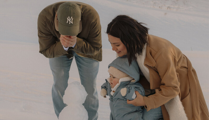 Candid family photo of a husband and wife building a mini snowman with their baby.