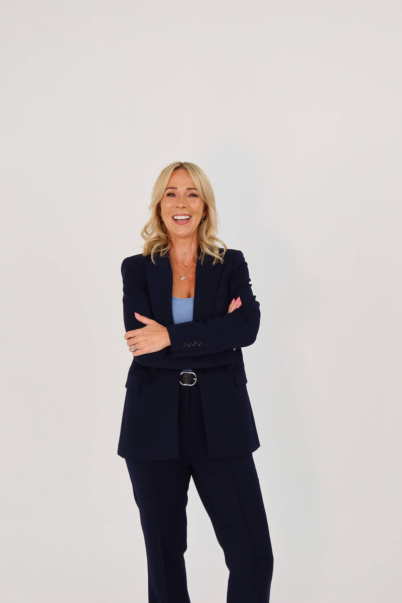 A woman with blonde hair, wearing a navy blue suit and a light blue top, stands against a plain white background with her arms crossed, smiling confidently.