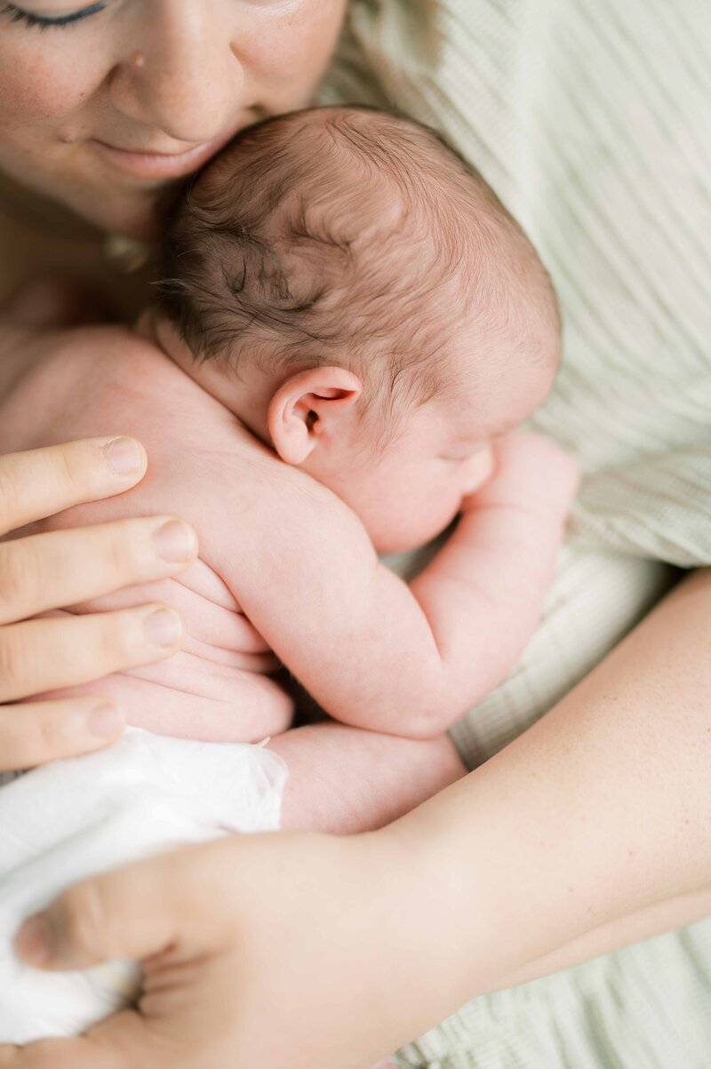 Woman wearing green floral sleeveless dress holds one year old son and kisses his hand as she looks out over the mountains in the distance by Portland Newborn Photographer Emilie Phillipson Photography.
