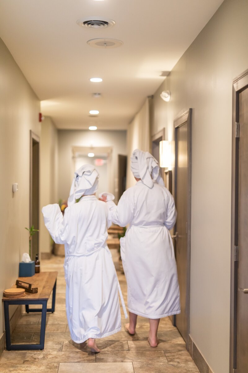 Two friends in robes and towels after spa services headed to sauna after dermaplaning and facials.