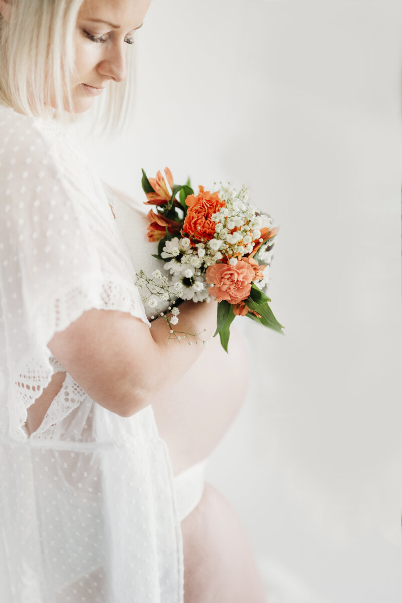 Studio maternity photo of a pregnant woman holding a bouquet of orange and white flowers against her bare belly, captured by a Kansas City maternity photographer.