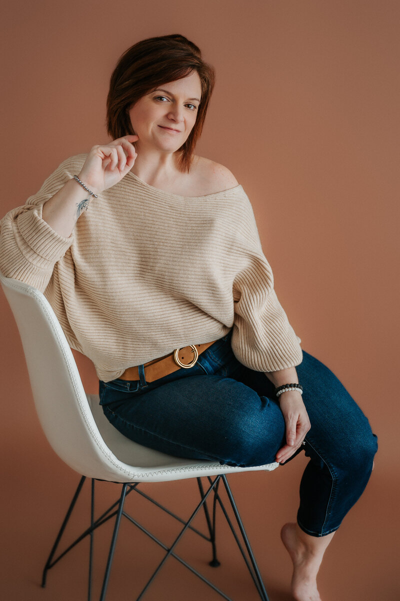 A woman with short brown hair sits sideways on a white chair against a brown background, wearing a beige sweater and blue jeans, with one hand raised near her face. Captured by a Macon GA Photographer.
