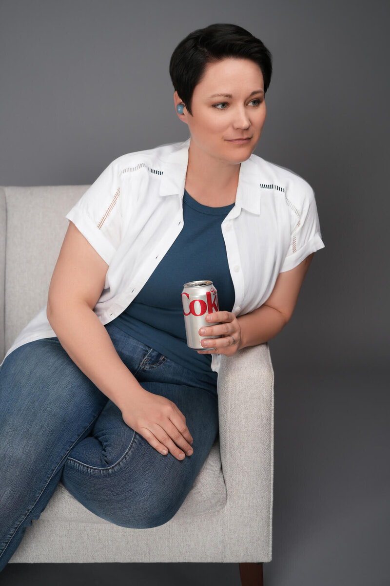 woman sitting in a chair holding a can of diet coke with an earbud in