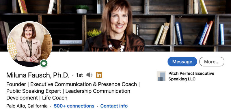 LinkedIn Profile Headshot with Banner Image Dr Miluna Fausch circle insert sitting with arms resting on crossed legs while smiling background photo cropped at head and shoulders smiling while standing in front of bookshelf